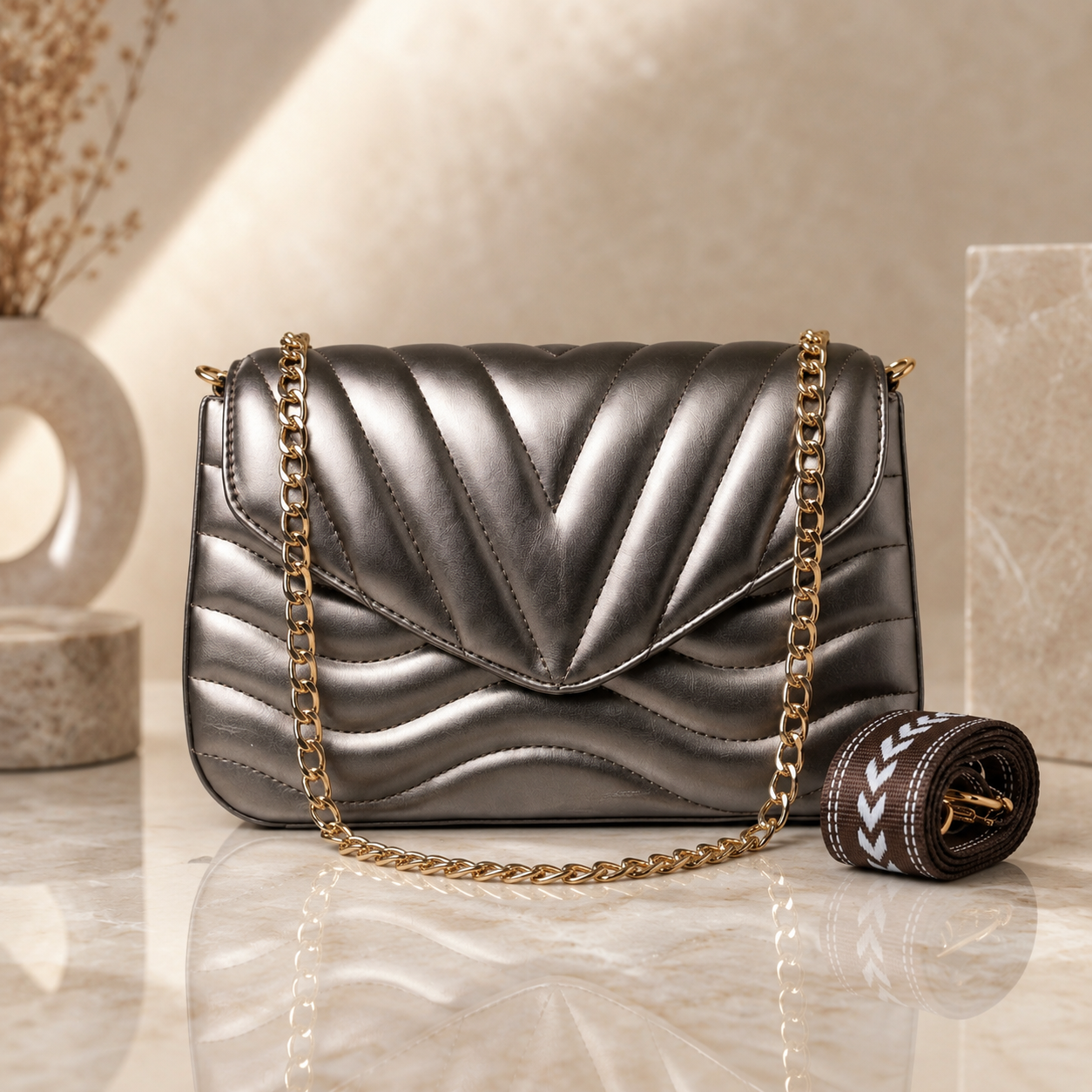 A metallic silver quilted flap shoulder bag with a gold chain strap, shown on a polished marble surface with a matching rolled brown strap beside it. The background features a warm beige textured wall and decorative stone props.